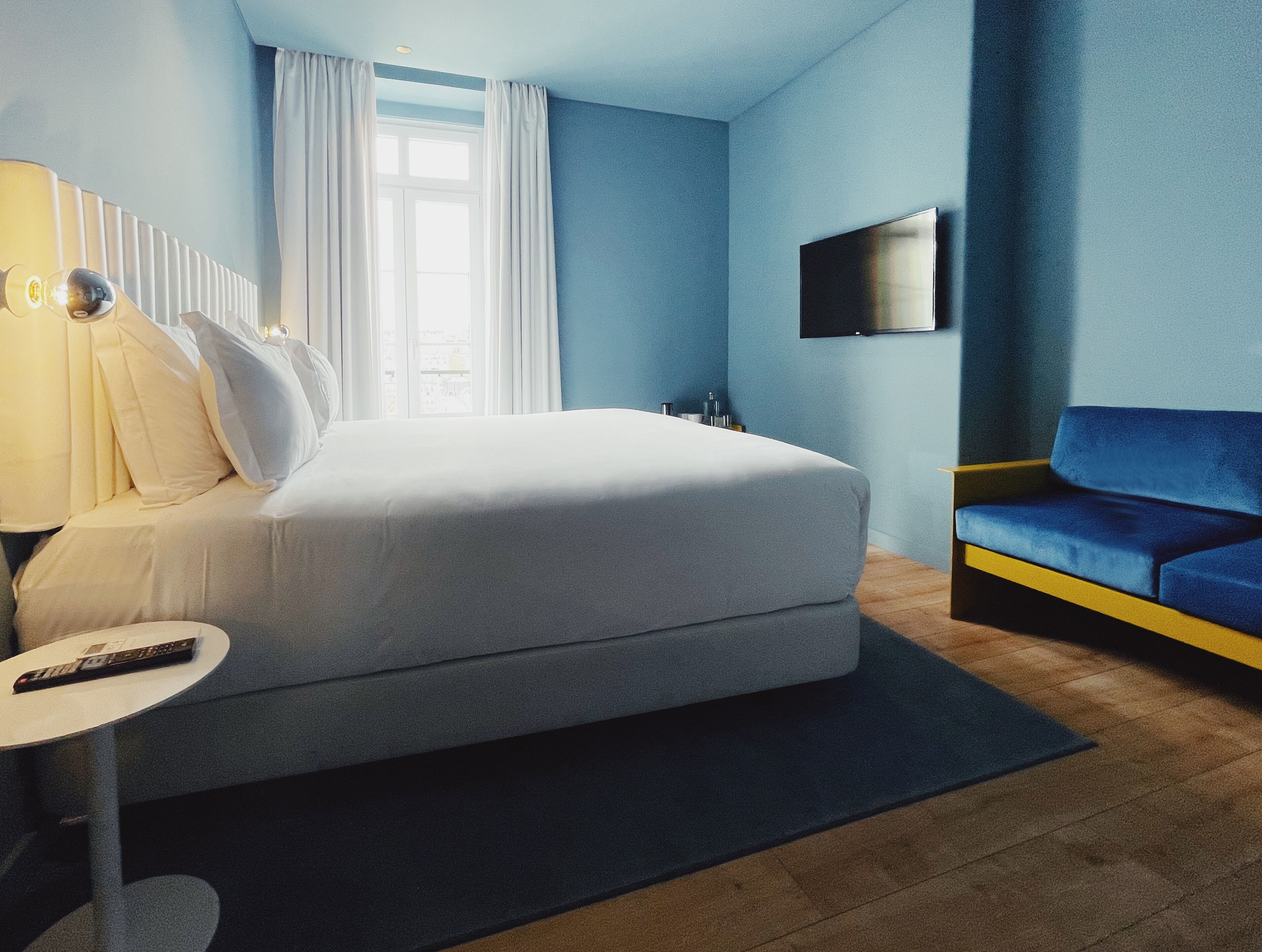 A modern hotel room with a large bed, a blue sofa, a TV on the wall, and a window with curtains.