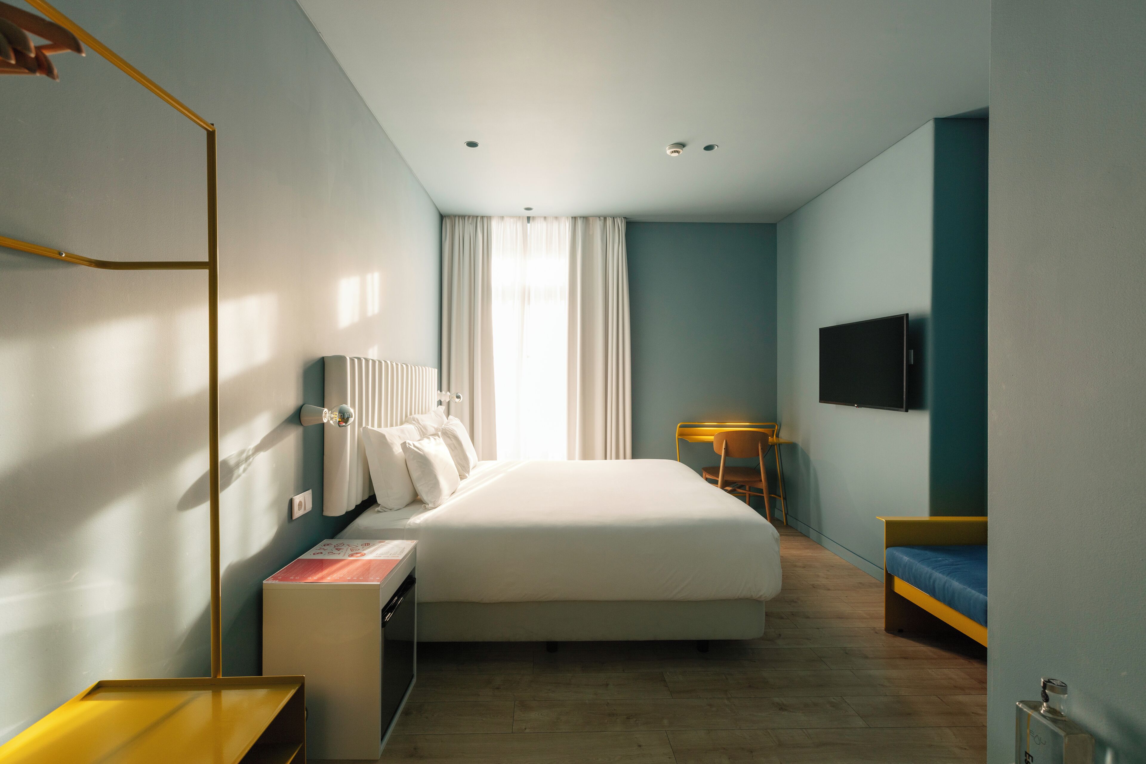 A modern hotel room with a large bed, a desk, a chair, a TV, and a window with curtains.