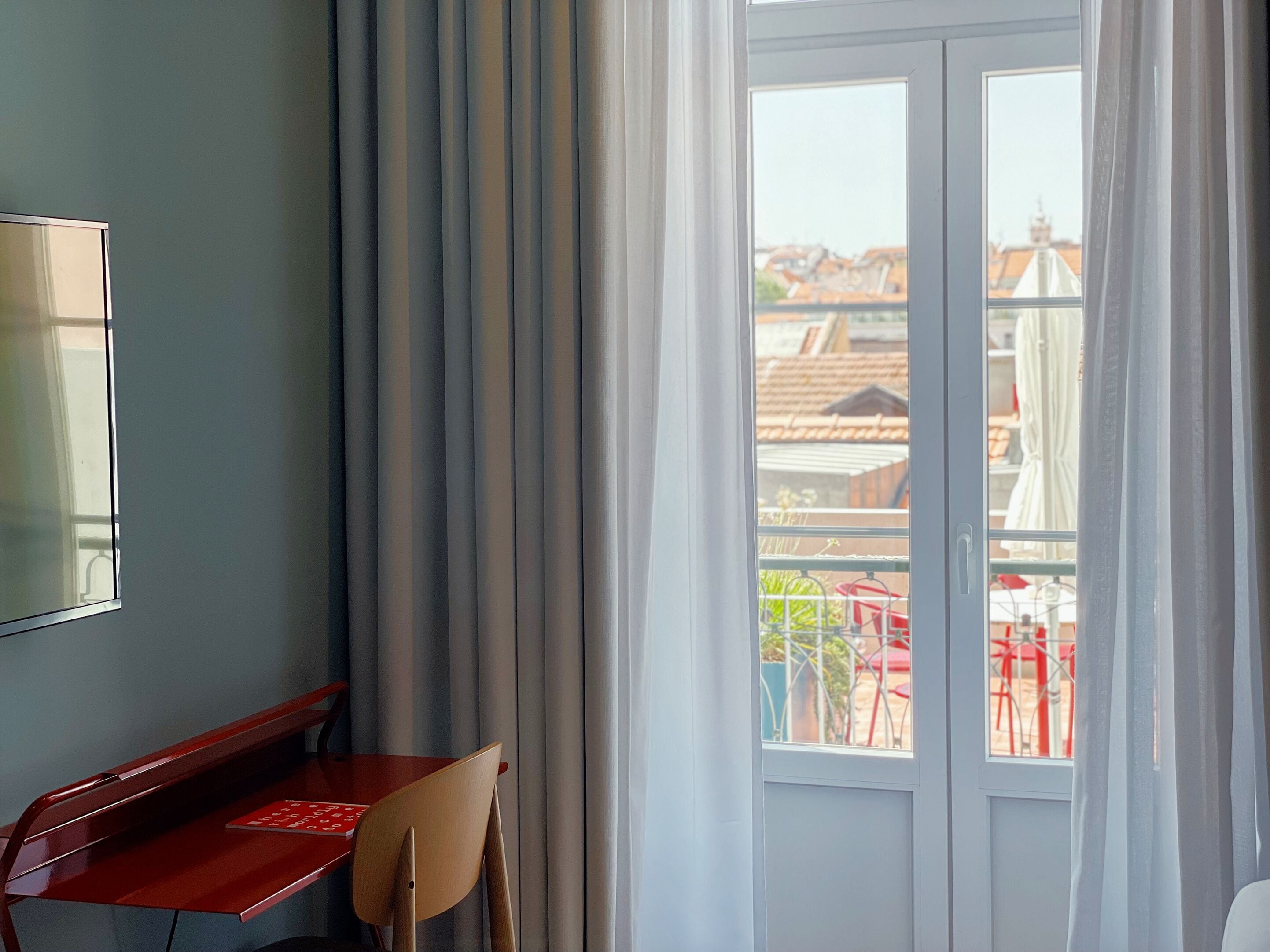 Een kamer met een groot raam, een houten stoel, een rode tafel en uitzicht op gebouwen en een balkon met rode stoelen.