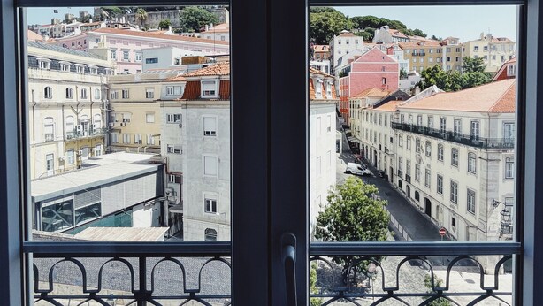 Una vista attraverso una finestra con ringhiera in metallo, che mostra una strada con edifici e cielo sereno.