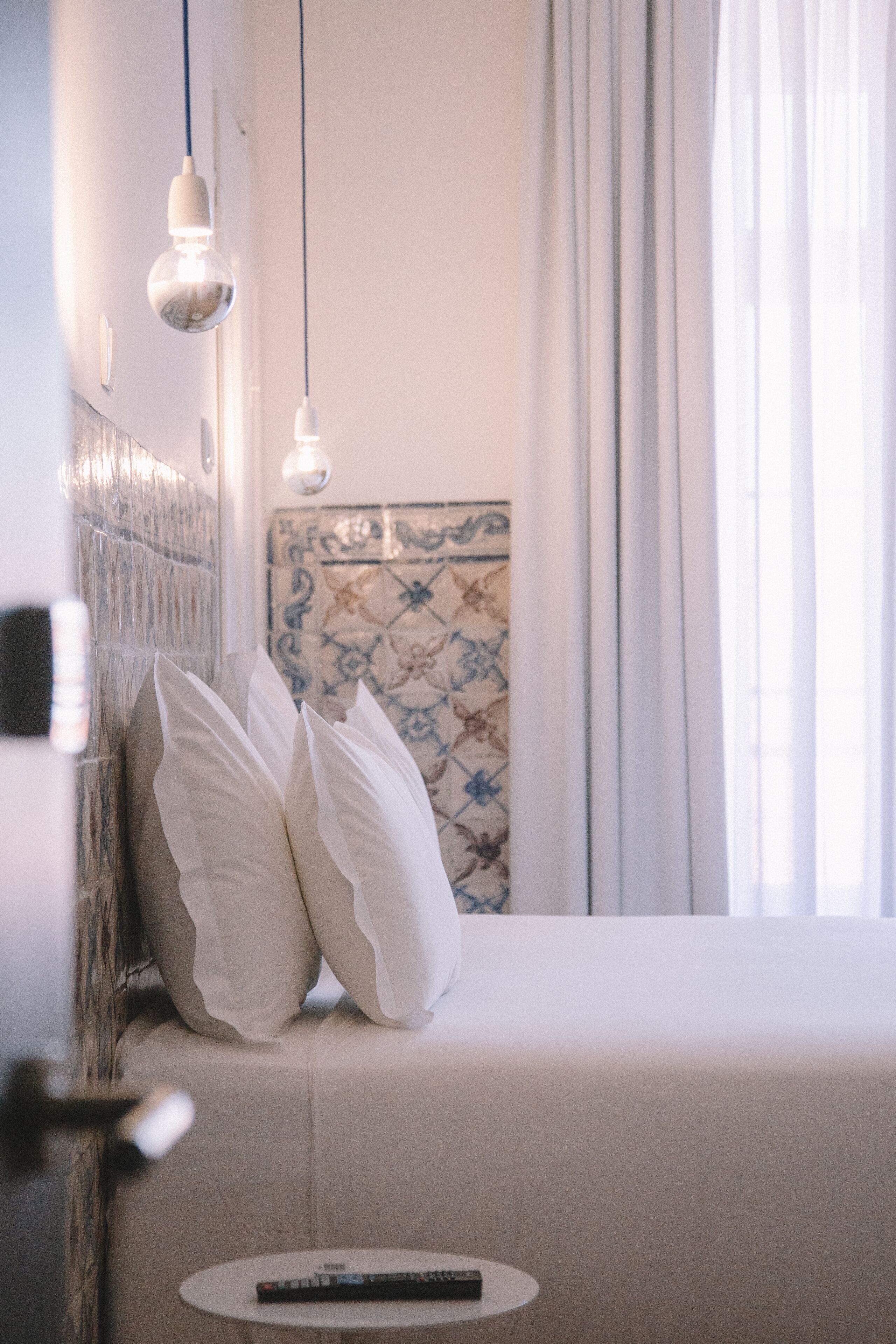 A neatly made bed with pillows, a remote control on a small table, and hanging light bulbs.