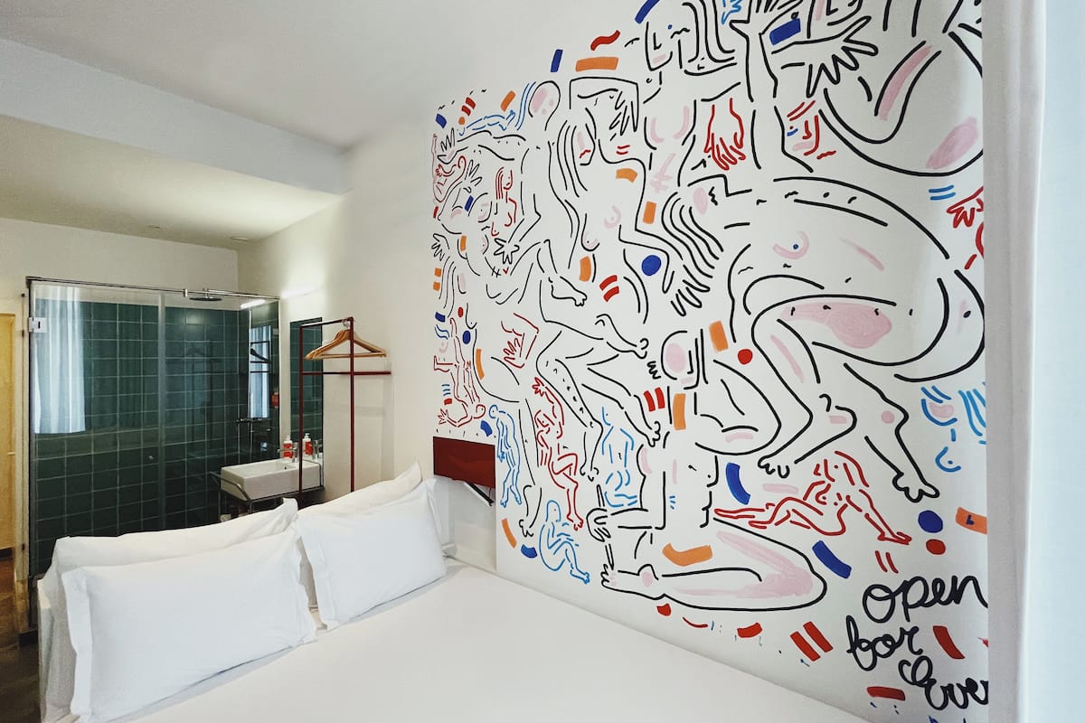 Een hotelkamer met een groot abstract schilderij op de muur, een bed met witte lakens, een badkamer met een glazen douchecabine en een hangende kapstok.
