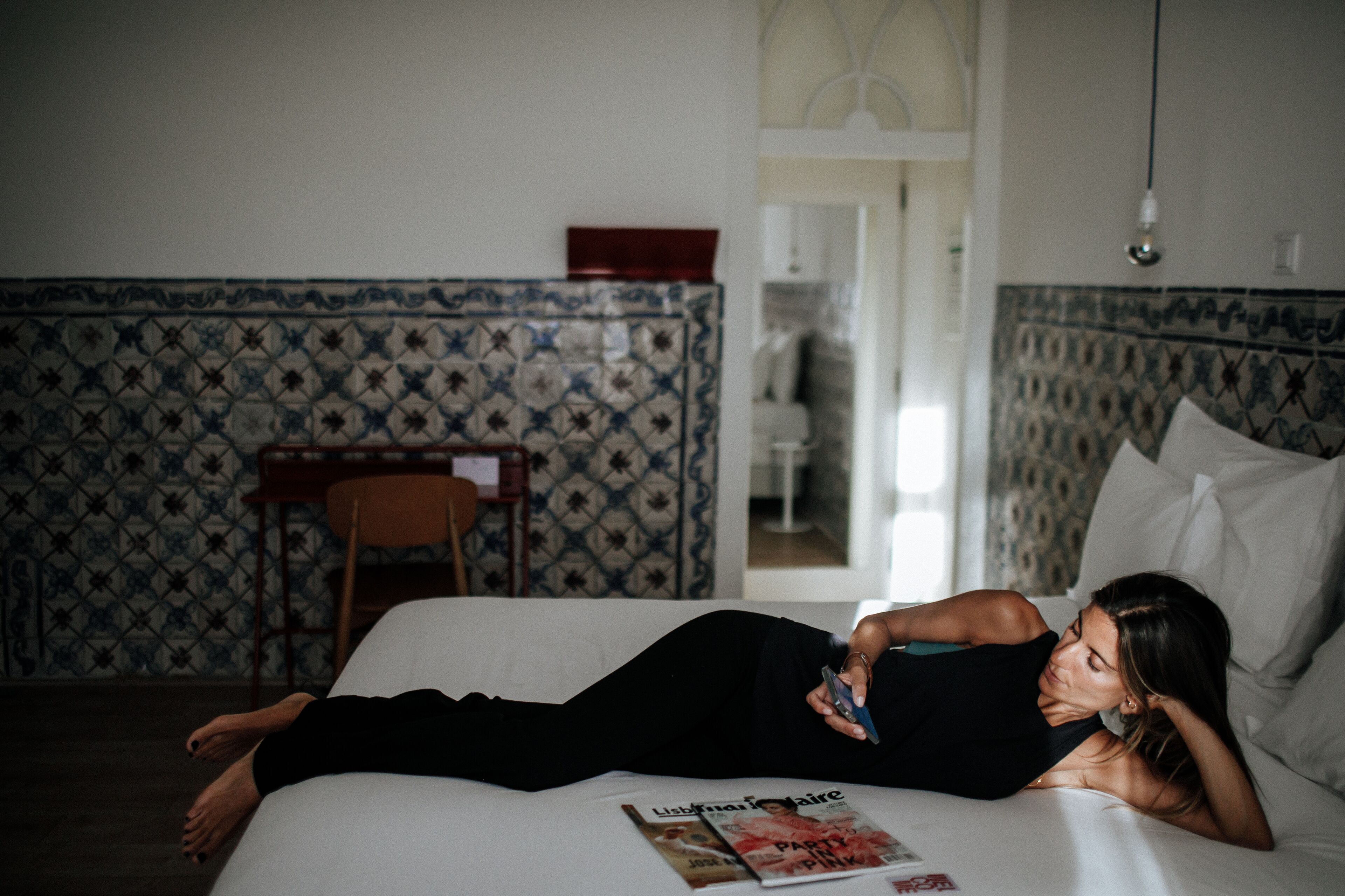A person lying on a bed in a room with patterned wallpaper and a visible magazine on the bed.