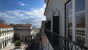 Vista da un balcone su una strada con edifici, cielo sereno e una distesa d'acqua in lontananza.