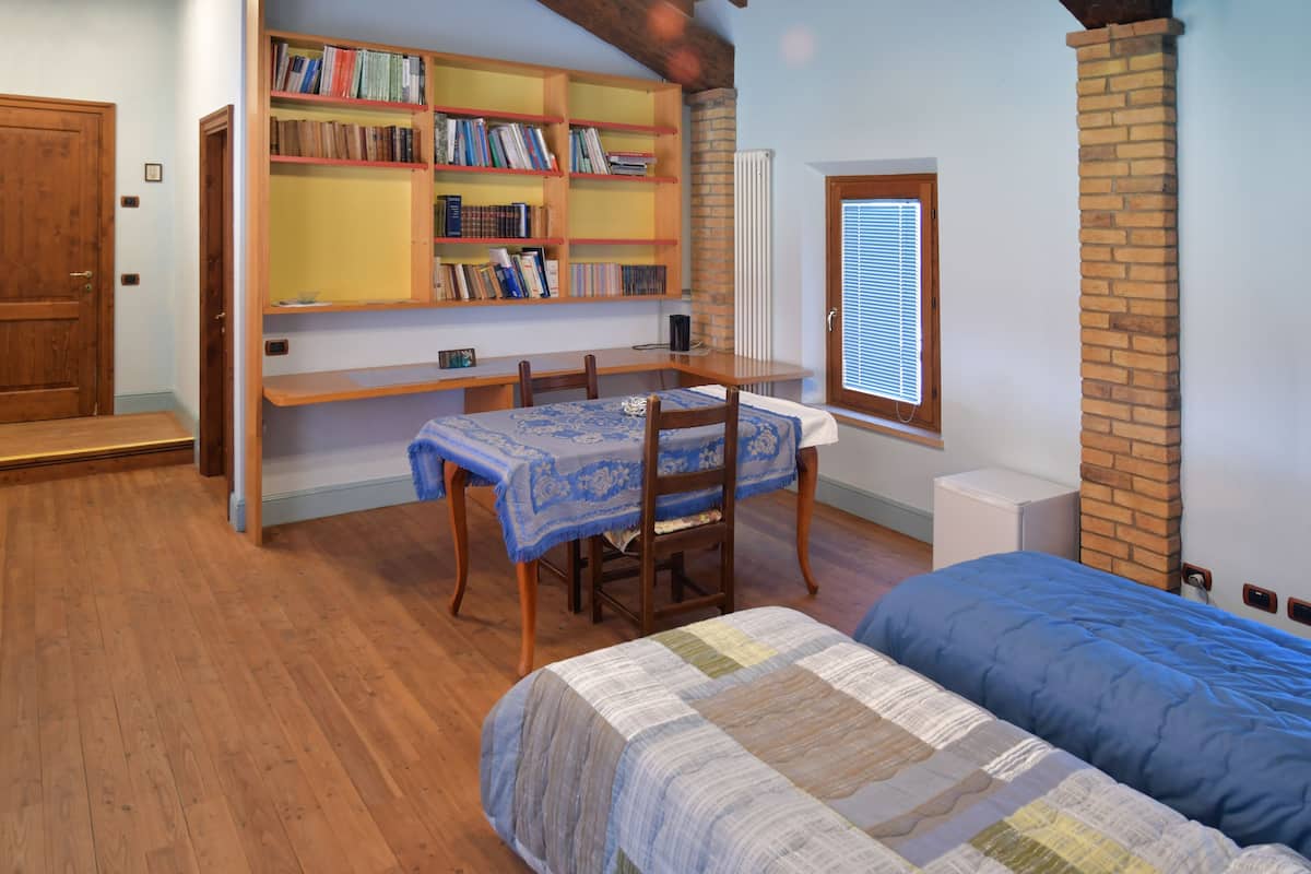Une pièce avec un plancher en bois, un lit recouvert d’une couette bleue et blanche, un bureau en bois avec une chaise, et une bibliothèque remplie de livres.