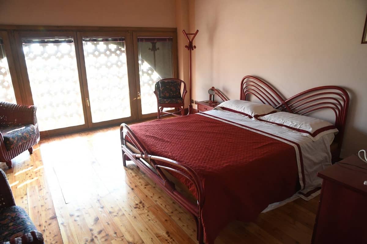 Une chambre à coucher avec un lit à ossature métallique, un plancher en bois, une chaise et une vue sur l’extérieur, visible à travers une porte vitrée.