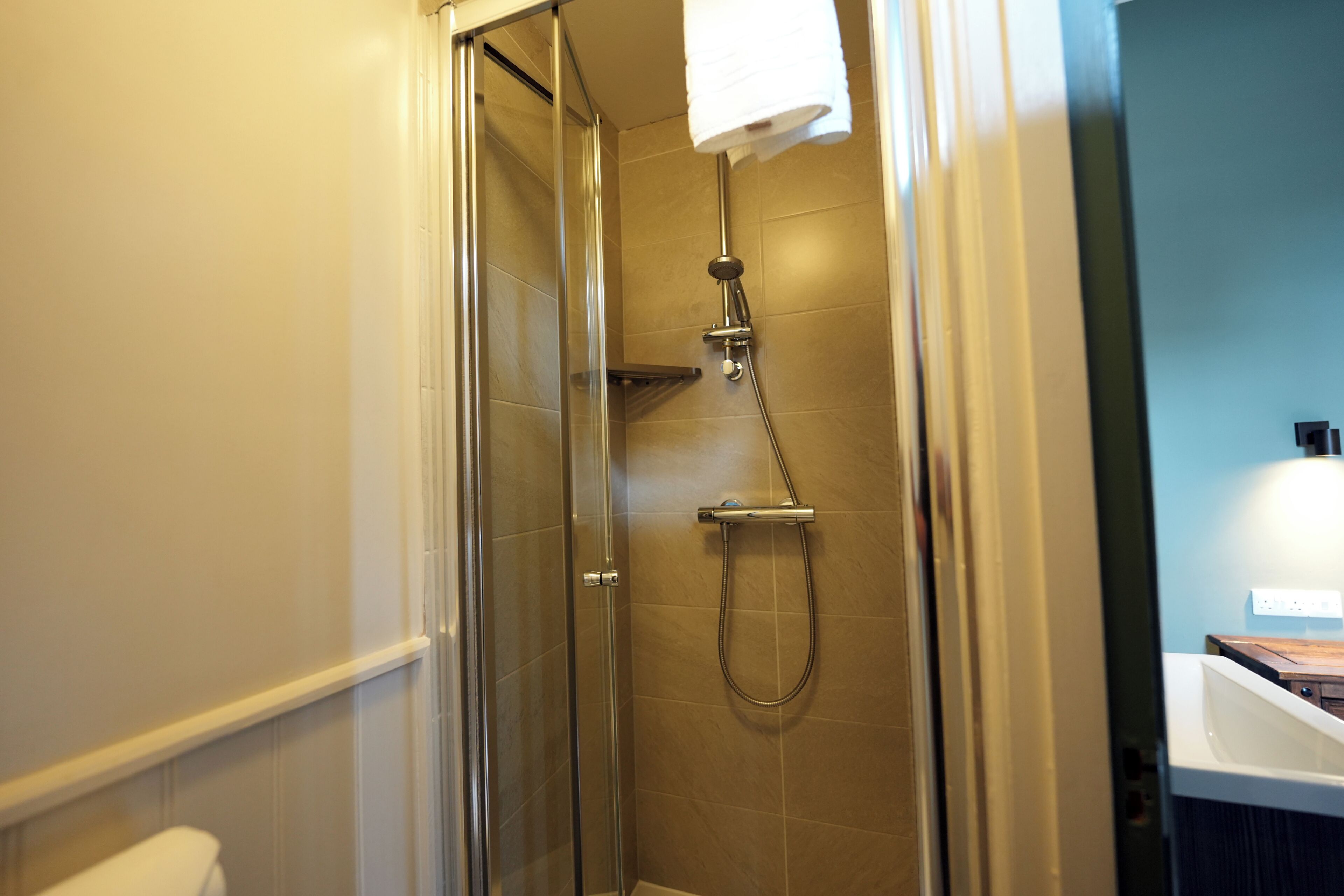 Ducha envuelta en vidrio con cabezal de ducha manual, una toalla colgada en la pared y un lavamanos blanco con encimera de madera.