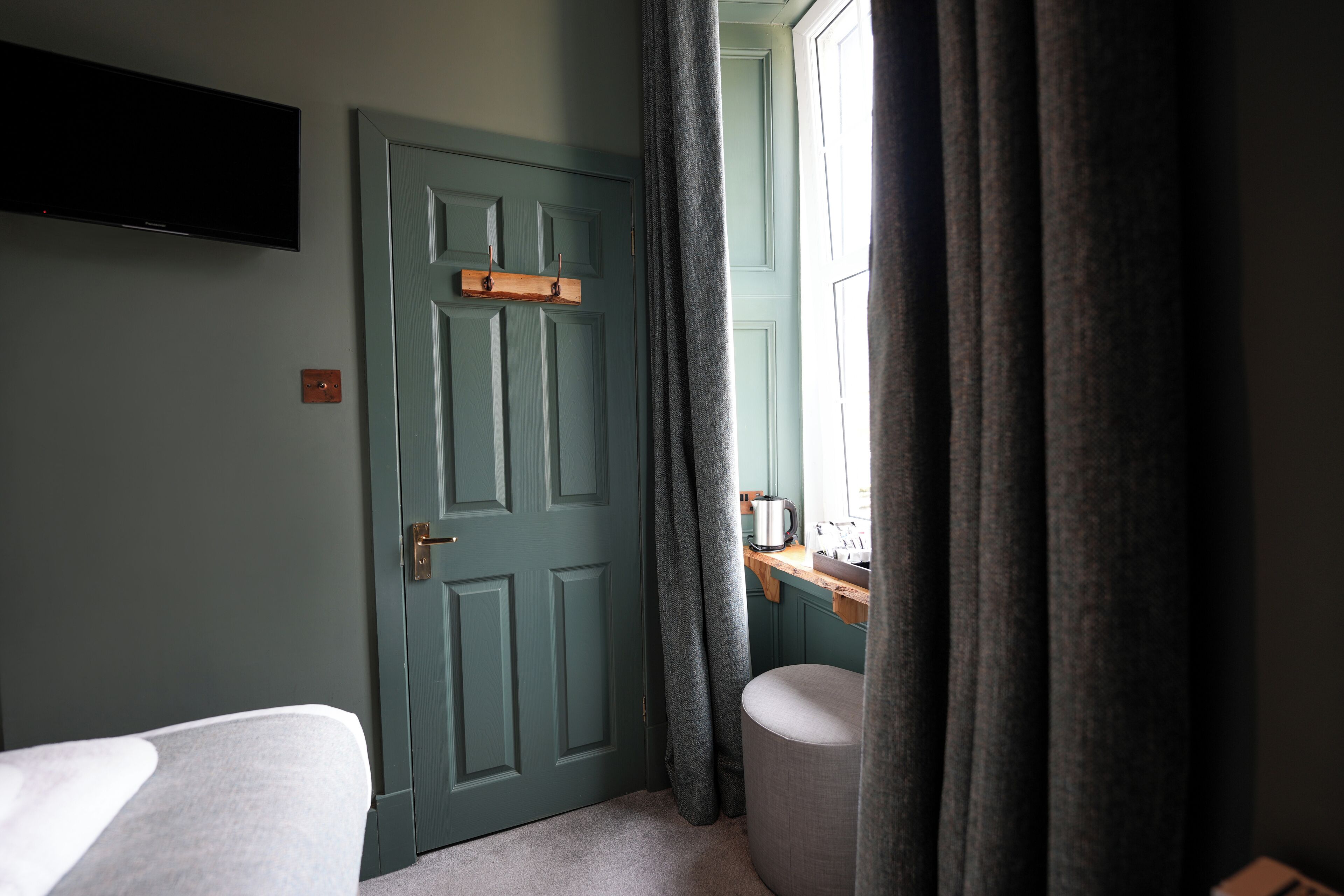 A room with a bed, a desk with a kettle, a window with curtains, and a door.
