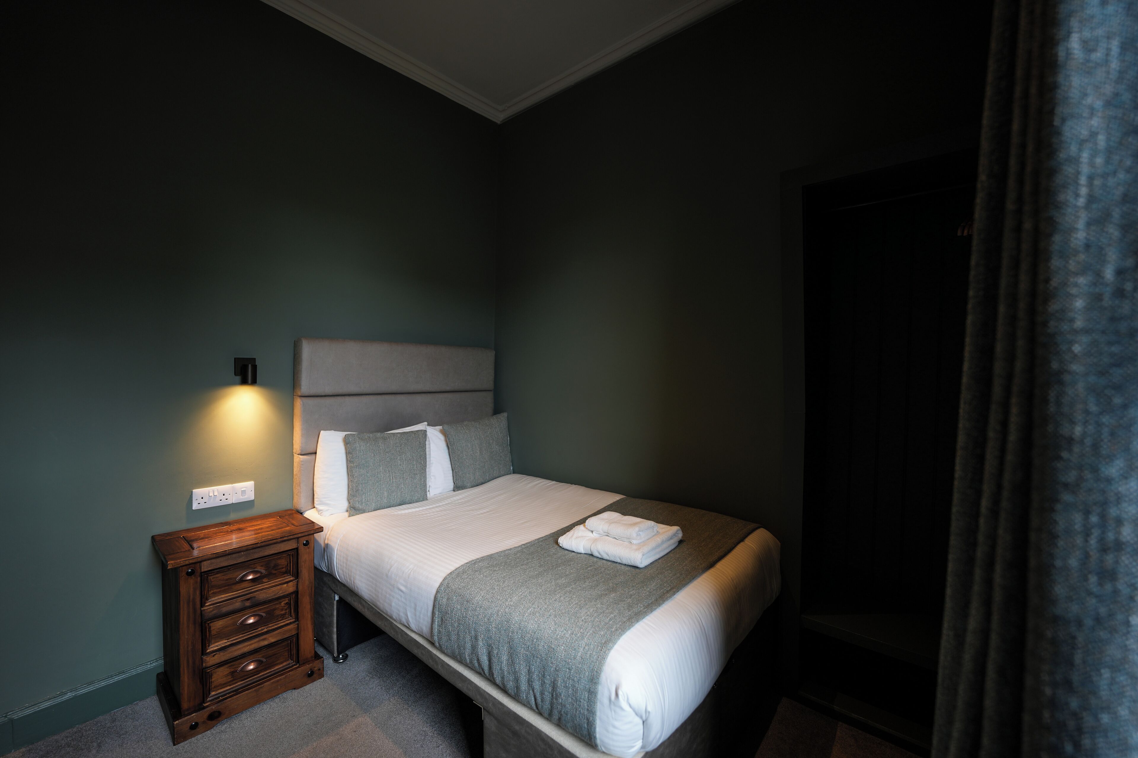 Una cama bien hecha con cabecera gris, una mesita de noche con cajones y un enchufe de pared.