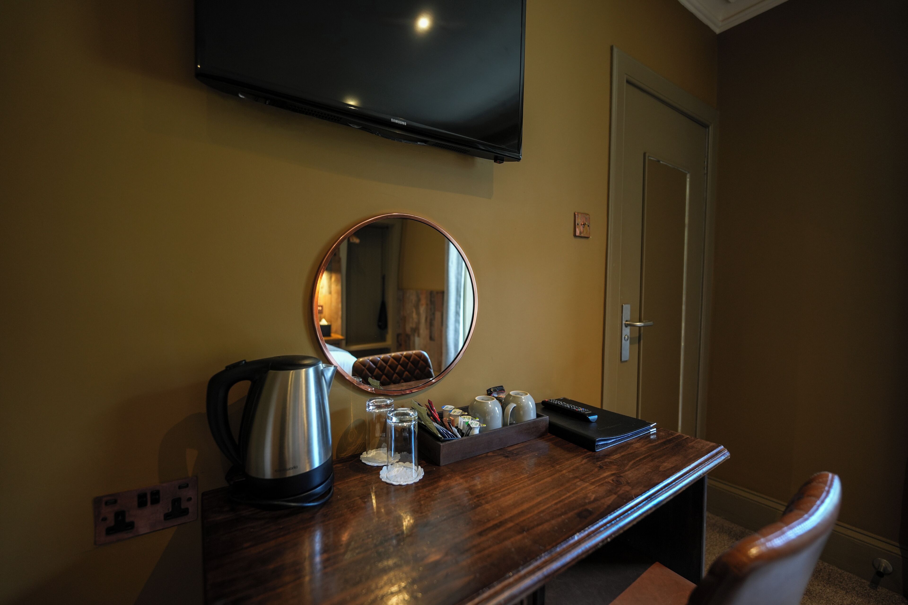 A hotel room with a wooden desk, a round mirror, a kettle, a tray with glasses and a remote control, and a television mounted on the wall.
