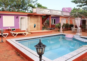 Ein Schwimmbecken mit Liegestühlen und Sonnensegel, umgeben von einem Gebäude mit violetten Fassade.