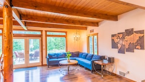 A spacious living room with a wooden ceiling, large windows, a blue sectional sofa, a small round table, and a world map on the wall.
