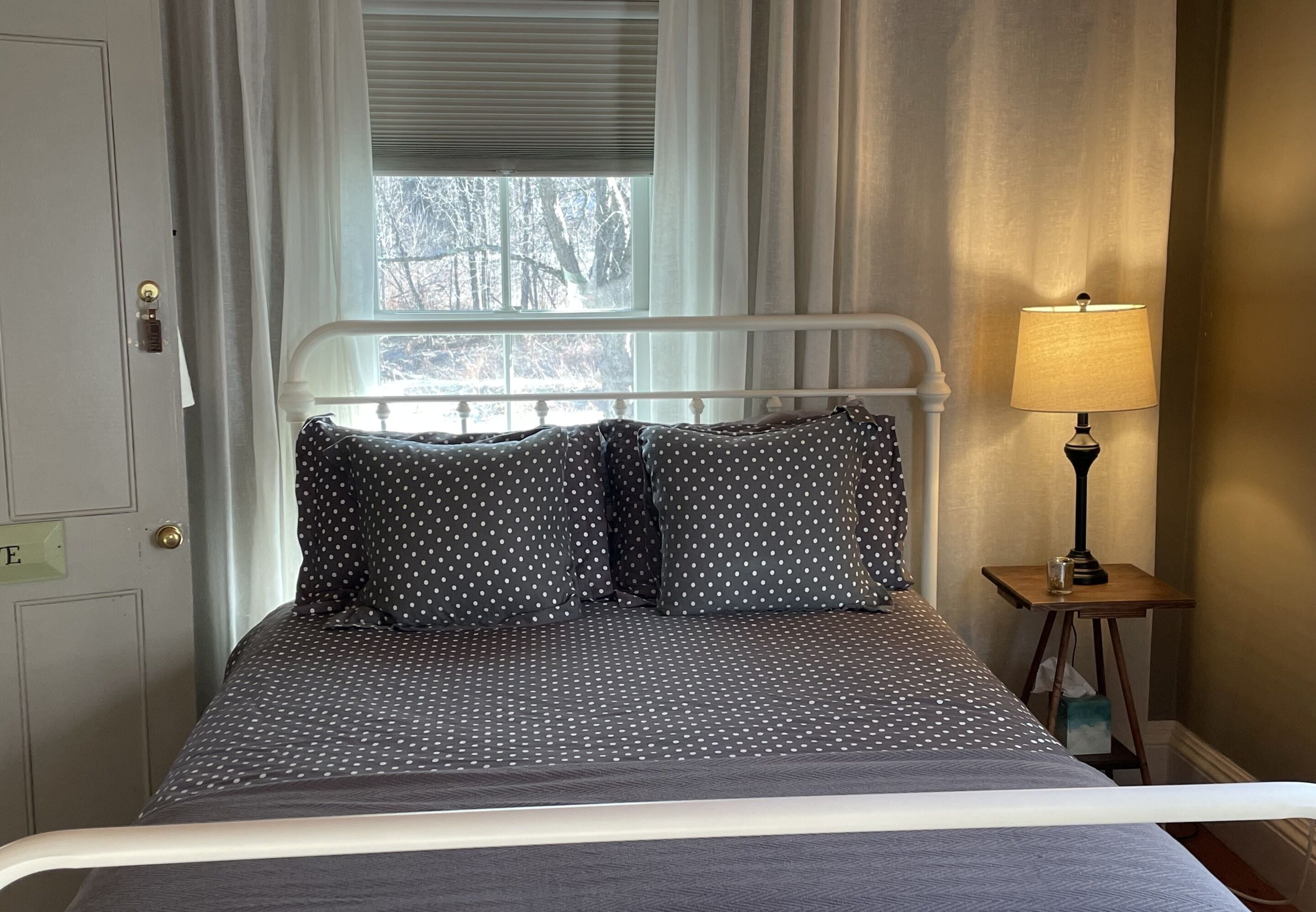 A bed with a white metal frame, dark polka-dotted bedding, and a bedside lamp on a small wooden table.