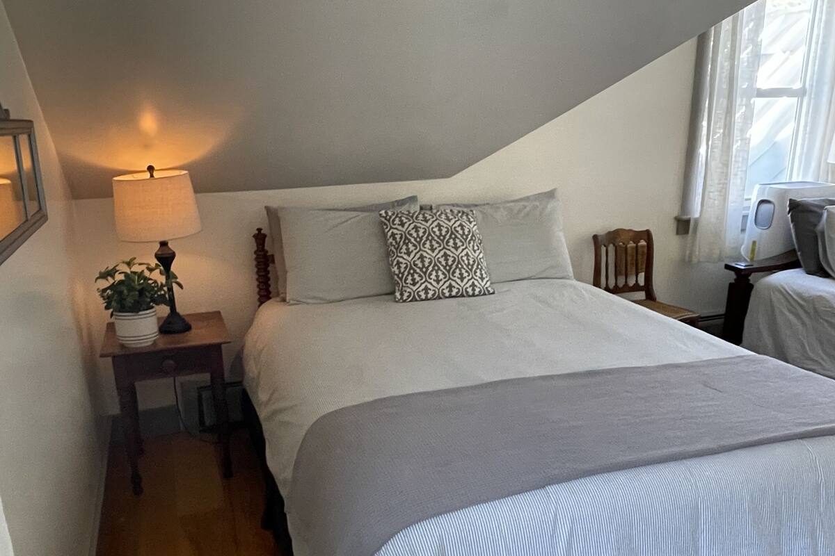 A bedroom with a bed, a bedside table with a lamp, a potted plant, and a window with curtains.