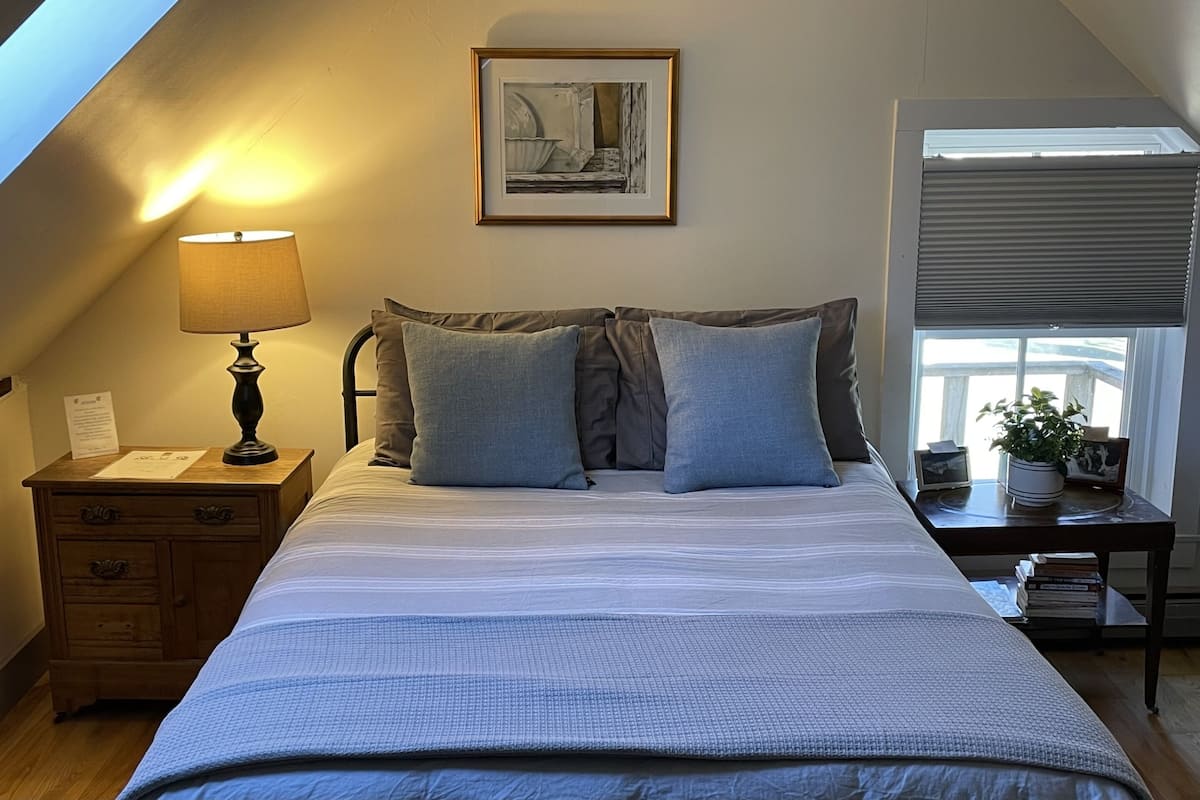 A bedroom with a bed, a nightstand, a lamp, a framed picture on the wall, a window with blinds, and a potted plant.
