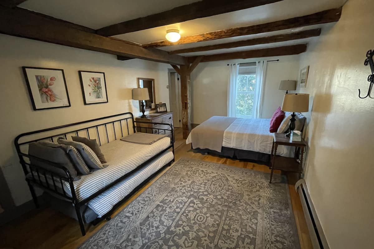 A bedroom with a single bed, a sofa bed, a patterned rug, wooden beams, and framed pictures on the wall.
