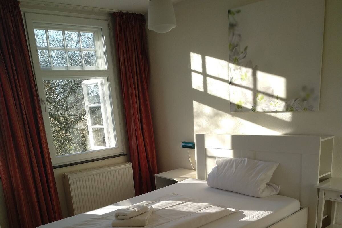 Un dormitorio con una cama grande, una mesita de noche, cortinas en una ventana y una lámpara colgante.