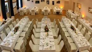Un salón de banquetes con varias mesas redondas, cada una con mantel blanco y sillas, y fotografías enmarcadas en las paredes.