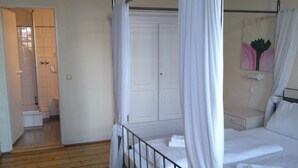 Una cama con dosel, ropa de cama blanca y cortinas, suelo de madera y una puerta blanca al fondo.