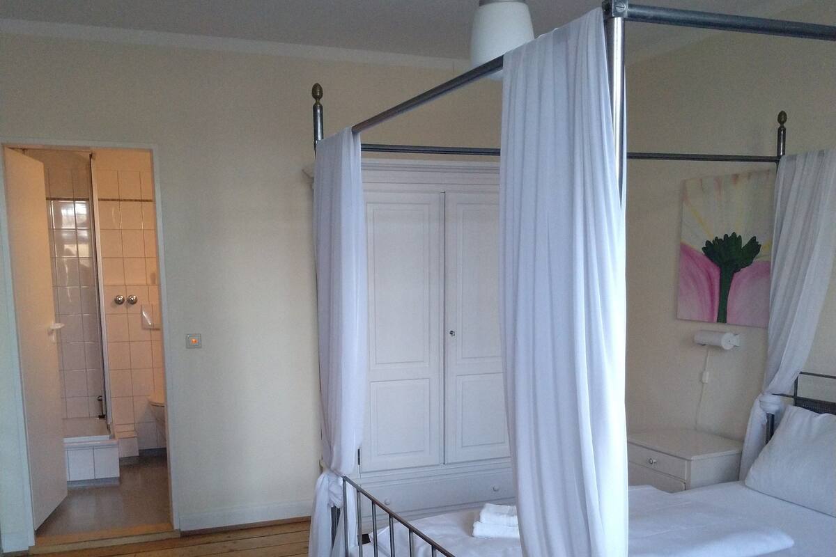 Una cama con dosel, ropa de cama blanca y cortinas, suelo de madera y una puerta blanca al fondo.