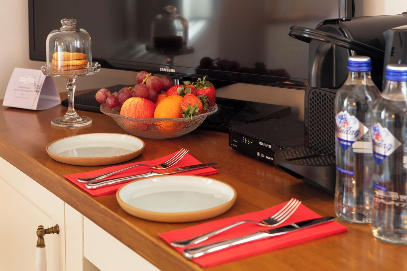 Habitación de hotel con zona de comedor que incluye un televisor, un bol de fruta, platos, cubiertos y agua embotellada.