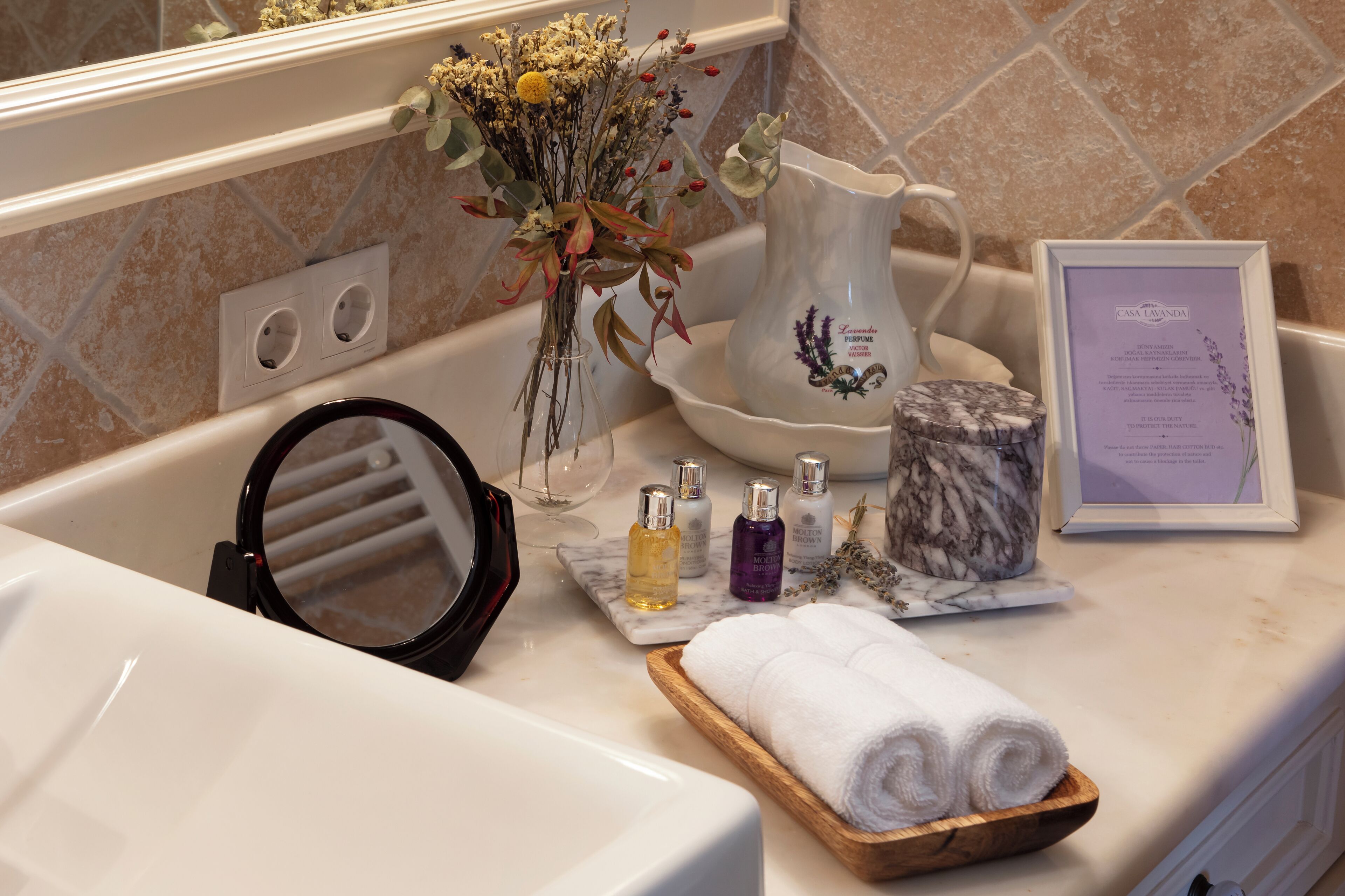 A bathroom with a marble sink, a towel rack, a vase with dried flowers, and a framed menu.