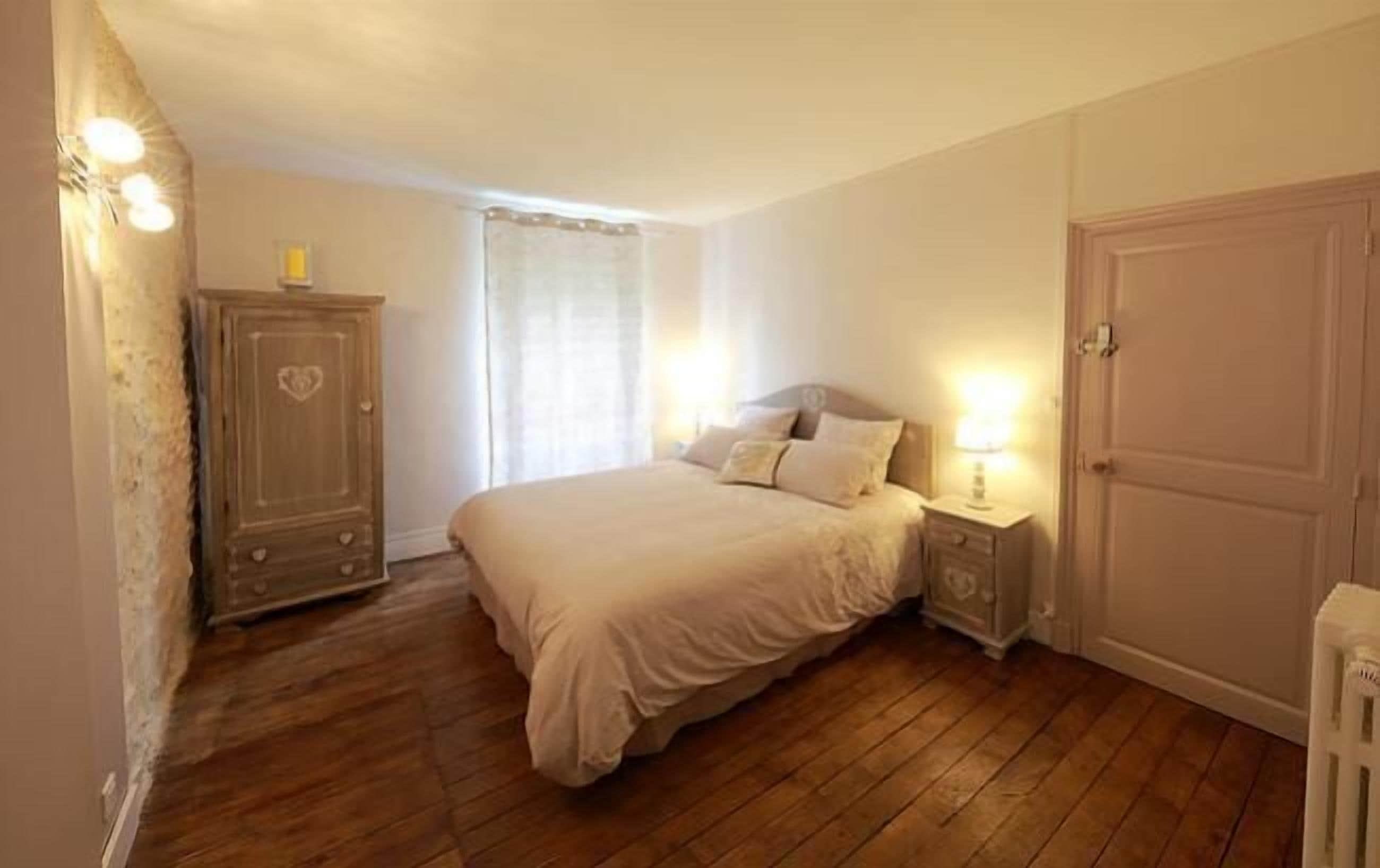A bedroom with a wooden floor, a bed with white bedding, a nightstand with a lamp, a wardrobe, and a door.