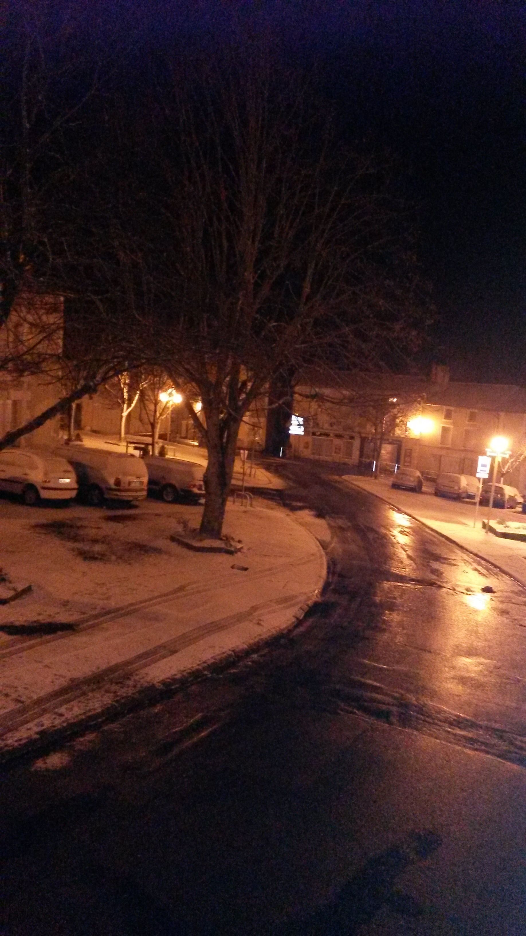 Una escena nocturna en la calle con coches aparcados y una acera cubierta de nieve.