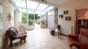 A spacious living room with a large glass door leading to a garden, a wooden bench, a brown leather armchair, and a bookshelf. - La Loge des Fées (Latille)