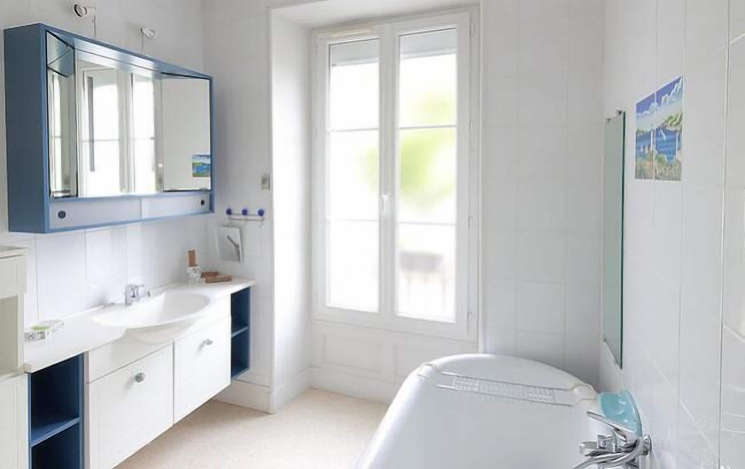 A modern bathroom with a large window, a white bathtub, and a blue-framed mirror.