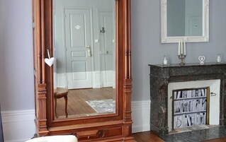 Um quarto com um espelho grande, uma lareira com estante de livros, e uma porta ao fundo.