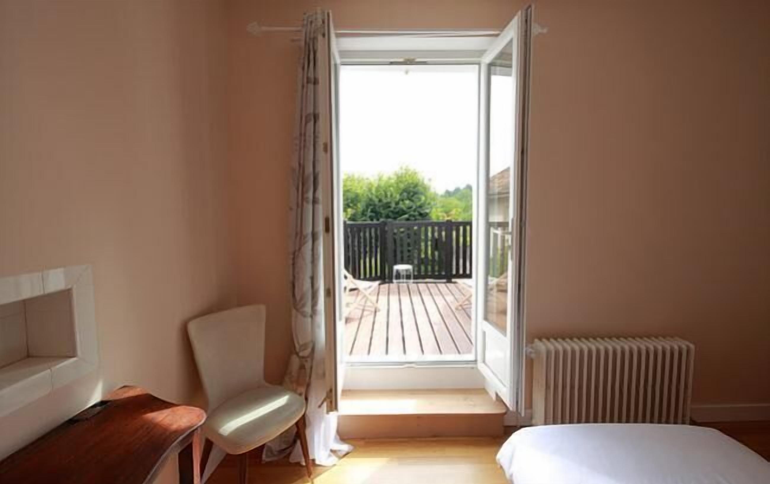 A bedroom with a bed, a chair, a small table, a radiator, and an open door leading to a balcony with a wooden deck and a black fence.