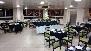 Une salle de banquet avec des tables rondes et des chaises, de grandes fenêtres offrant une vue sur la ville et une scène centrale.