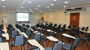 Une salle de conférence avec des rangées de chaises et de tables, un tableau blanc et un écran de projection.