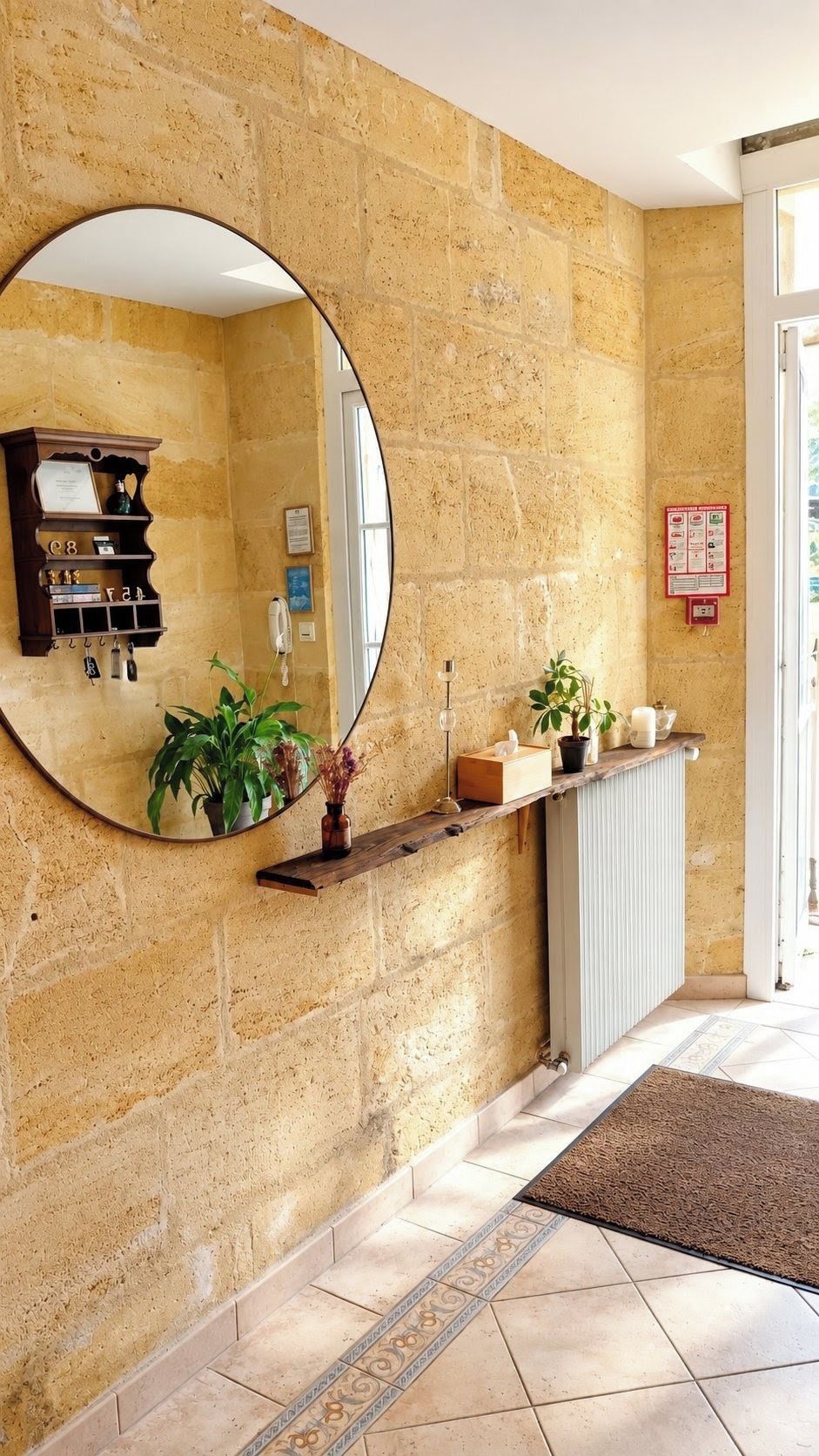 Un hall con paredes de piedra, un espejo grande, una estantería de madera con plantas, un radiador y un suelo de baldosas.