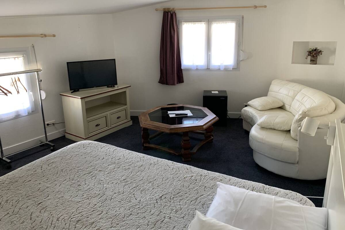 Ein Hotelzimmer mit Bett, Fernseher auf einem Schrank, einem kleinen Tisch, einer Couch und einem Fenster mit Vorhängen.