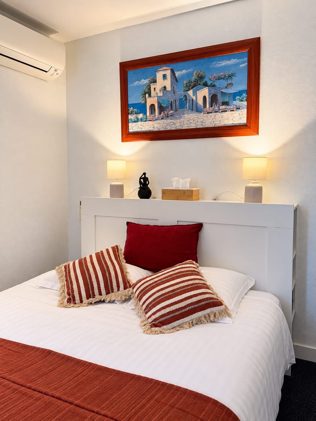 Una habitación de hotel con una cama, dos almohadas, una mesita de noche, una lámpara, un cuadro y un aire acondicionado.