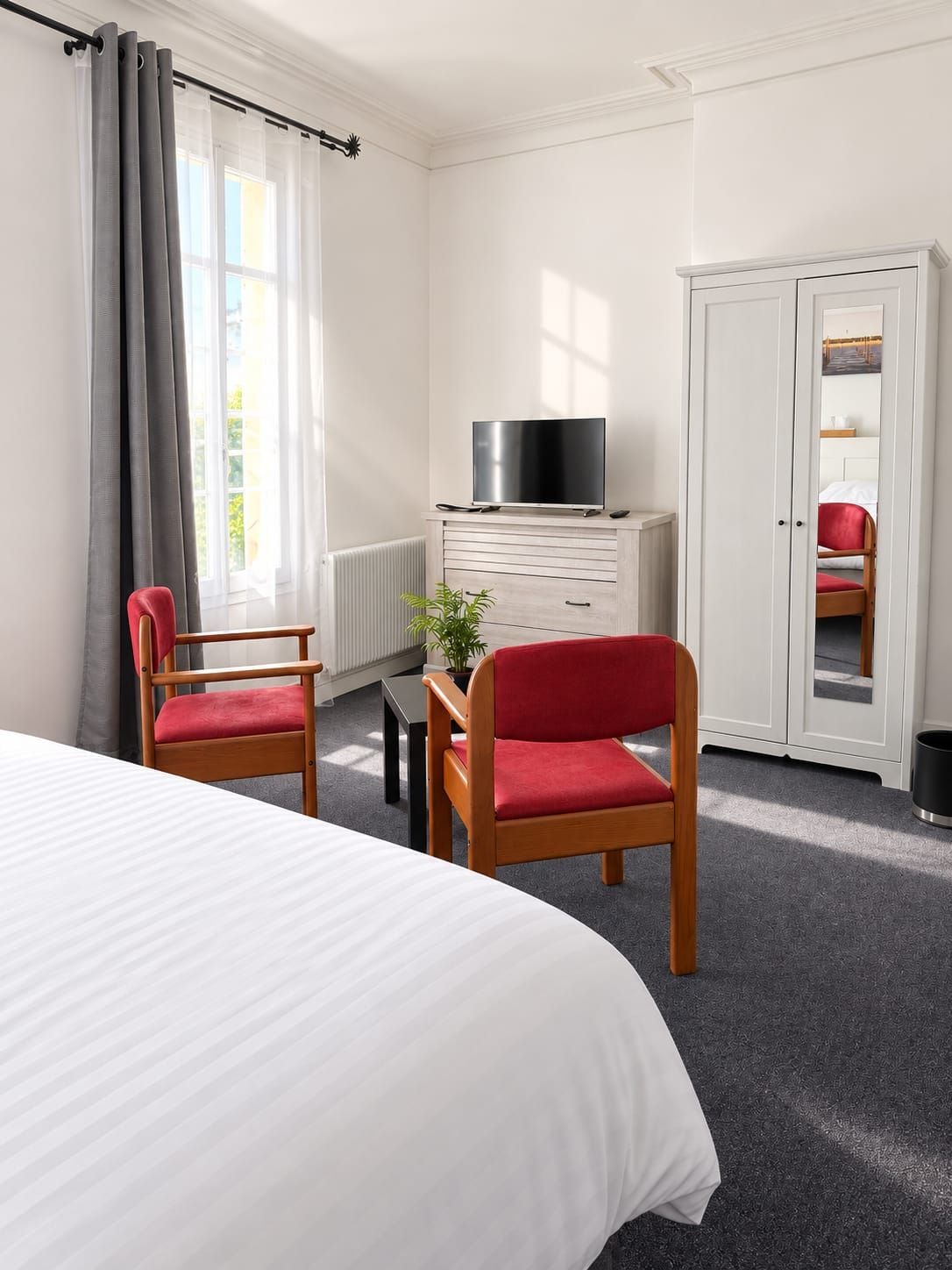 Quarto de hotel com uma cama, duas cadeiras, uma televisão sobre um aparador e um guarda-roupa.
