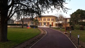 A pathway leading to a building with a tiled roof and red shutters. There is a small play area with a colorful structure and a bench.