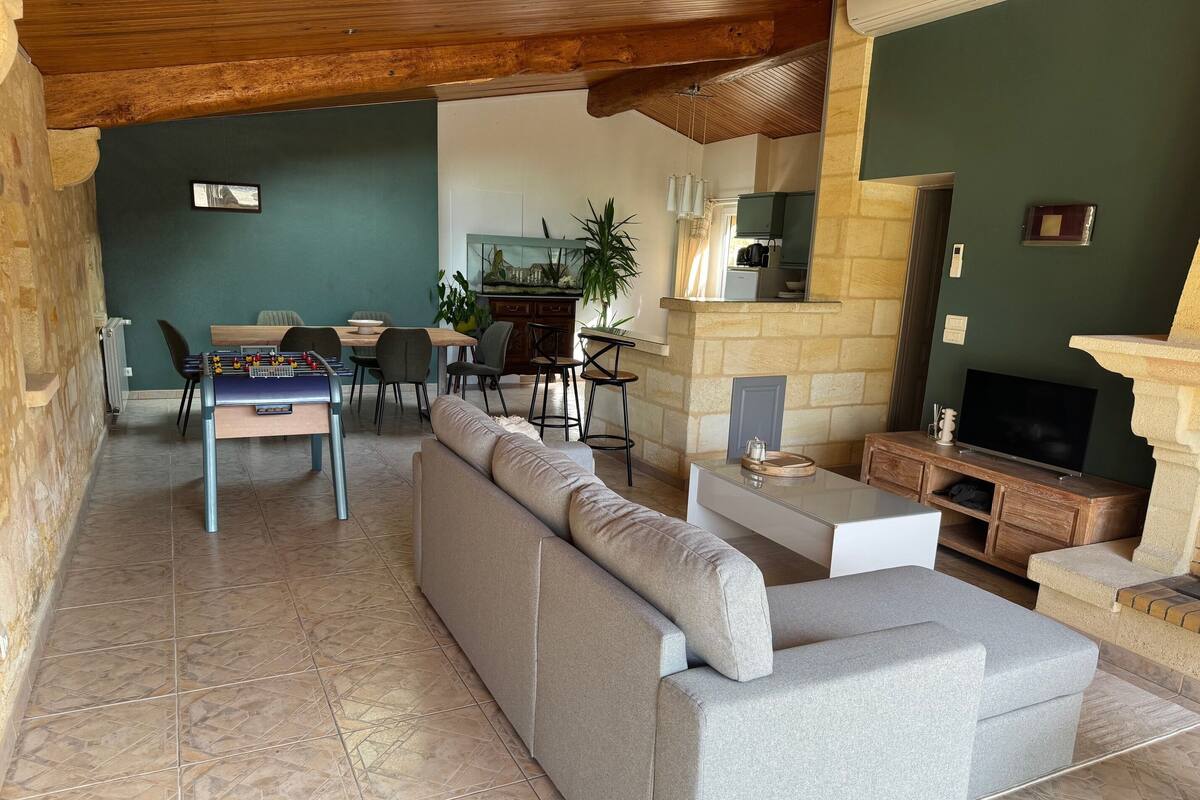 Ein Wohnzimmer mit einer Couch, einem Kicker, einem Essbereich mit Stühlen, einer Küche im Hintergrund und einem Steinkamin.