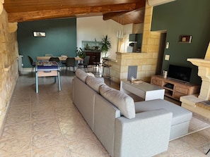 A living room with a sofa, a foosball table, a dining area with chairs, a kitchen in the background, and a stone fireplace. - Le Melchior (Sainte-Terre)