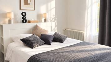 A bedroom with a bed, pillows, a bedside table with lamps, a radiator, and a window with curtains.