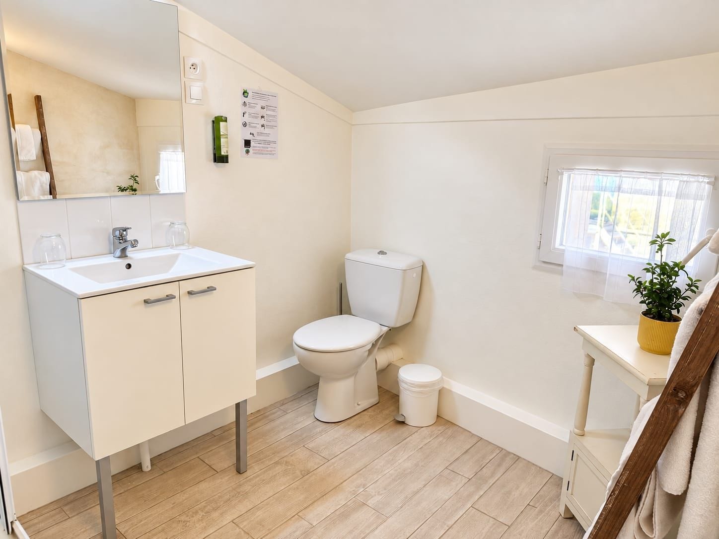 A bathroom with a white sink, toilet, and bidet, wooden flooring, and a window with a plant.