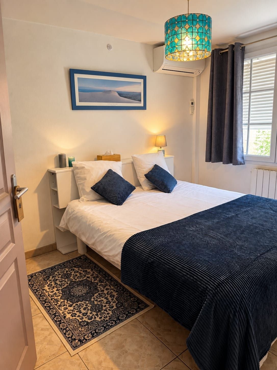 A bedroom with a bed, a nightstand, a lamp, a picture on the wall, and a window with curtains.