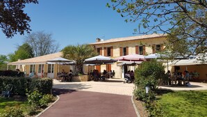 An outdoor café area with tables and umbrellas, surrounded by greenery and a building with a tiled roof.