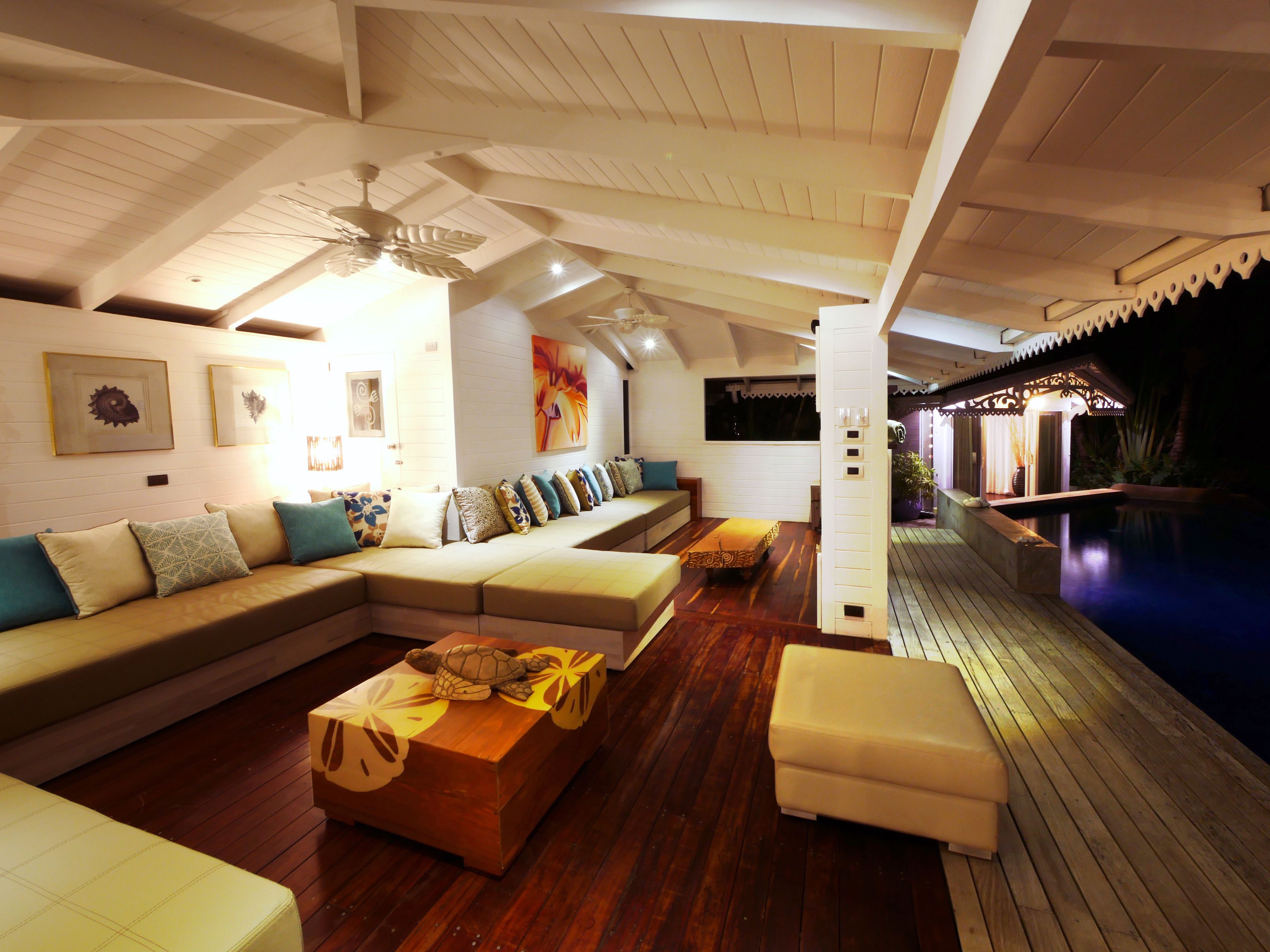 A spacious living area with a wooden floor, a large sectional sofa, a wooden coffee table, and a view of an outdoor swimming pool at night.