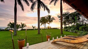 A wooden deck with a curved bench, surrounded by palm trees and a grassy area, overlooking a body of water. - Villa Diosa del Mar (Veintisiete de Abril)