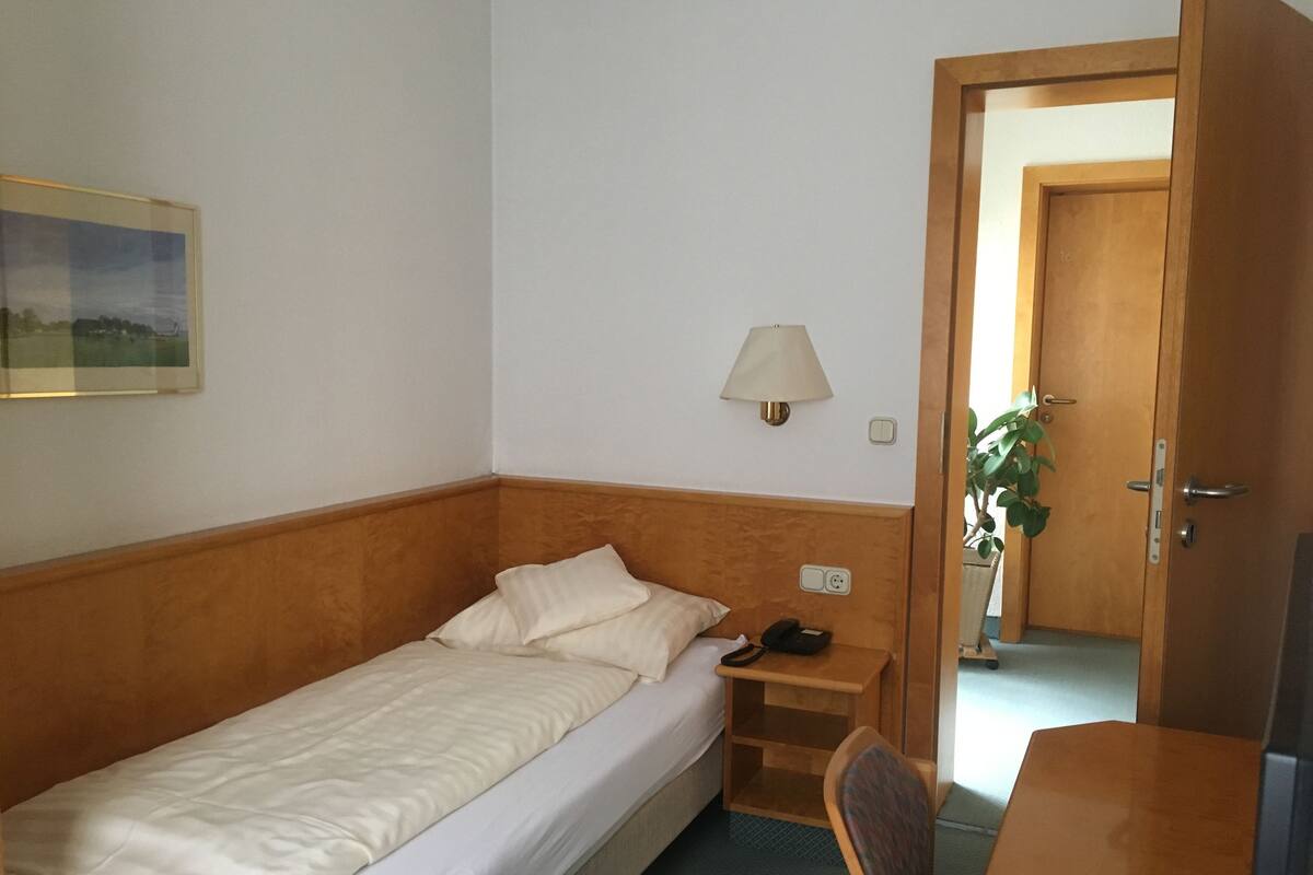 Ein Einzelbett mit Holz Kopfteil, ein Nachttisch mit Lampe, eine kleine Pflanze und eine Tür zu einem anderen Raum.