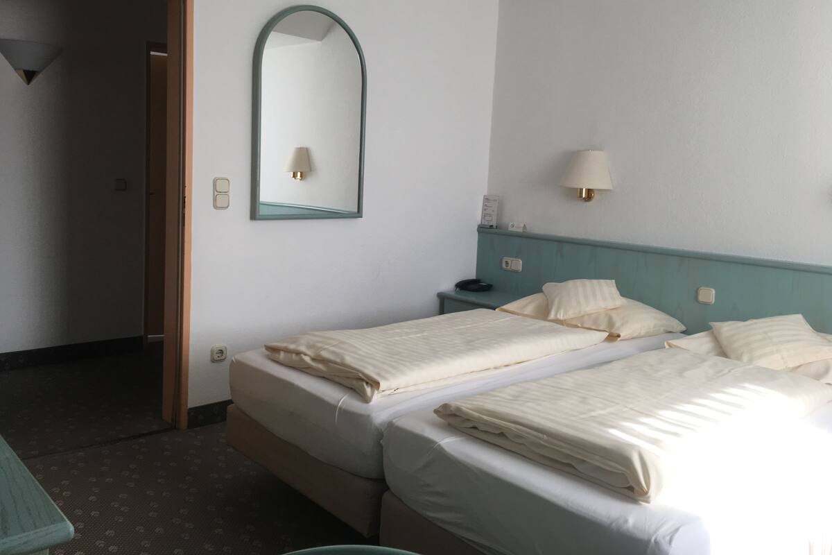 Ein Hotelzimmer mit zwei Betten, einem Spiegel, Nachttischlampen und einer Tür zu einem anderen Raum.