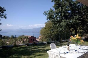 Un espace de restauration en plein air avec une table dressée pour deux personnes, offrant une vue panoramique sur un plan d’eau et des montagnes.